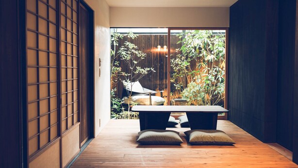 Ein traditioneller japanischer Raum mit Tatami-Boden, einem niedrigen Tisch, Kissen und einer Schiebetür, die zu einem Garten führt.