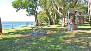 Relax in a serene park setting with a tranquil water view, surrounded by mature trees and equipped with picnic tables and a covered seating area.