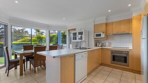Enjoy meals in the adjacent dining area with a view of the surrounding greenery, adjacent to a well-equipped kitchen with a dishwasher, oven, and microwave.