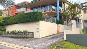 Ein modernes Wohnhaus mit zwei Etagen, einem Balkon, einer mit Pflanzen bepflanzten Steinmauer und einem gepflasterten Einfahrtsweg.