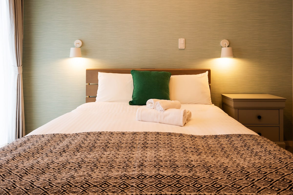 Un lit bien fait, recouvert d’une couvre-lit à motifs, avec deux oreillers et un coussin décoratif vert. On y trouve deux lampes de chevet et une table de chevet dotée d’un tiroir.