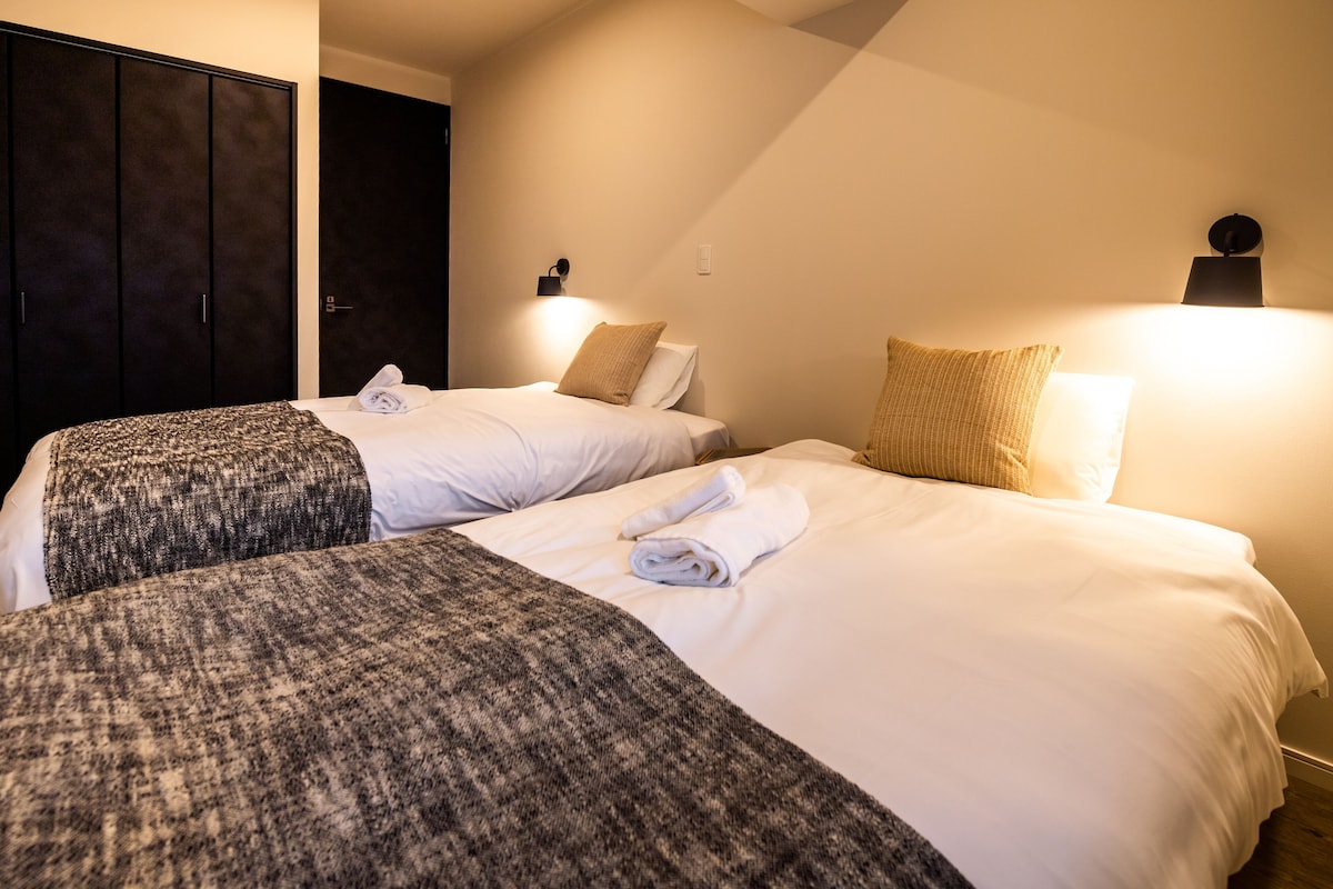 Une chambre d’hôtel avec deux lits, chacun recouvert d’une couverture grise et de draps blancs. Deux lampes murales éclairent la pièce.
