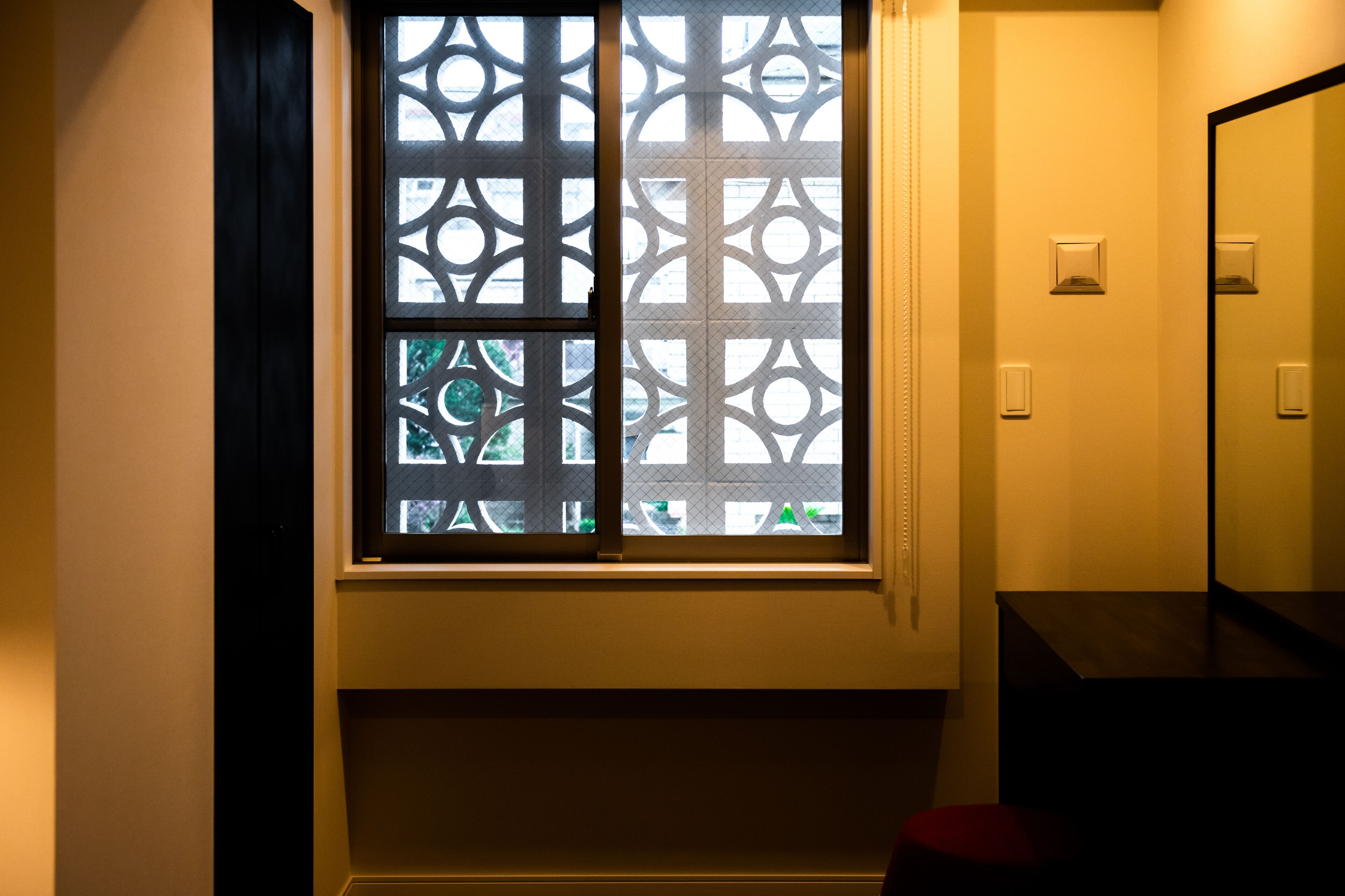 A bathroom with a window featuring geometric patterns, a mirror, and a red stool.