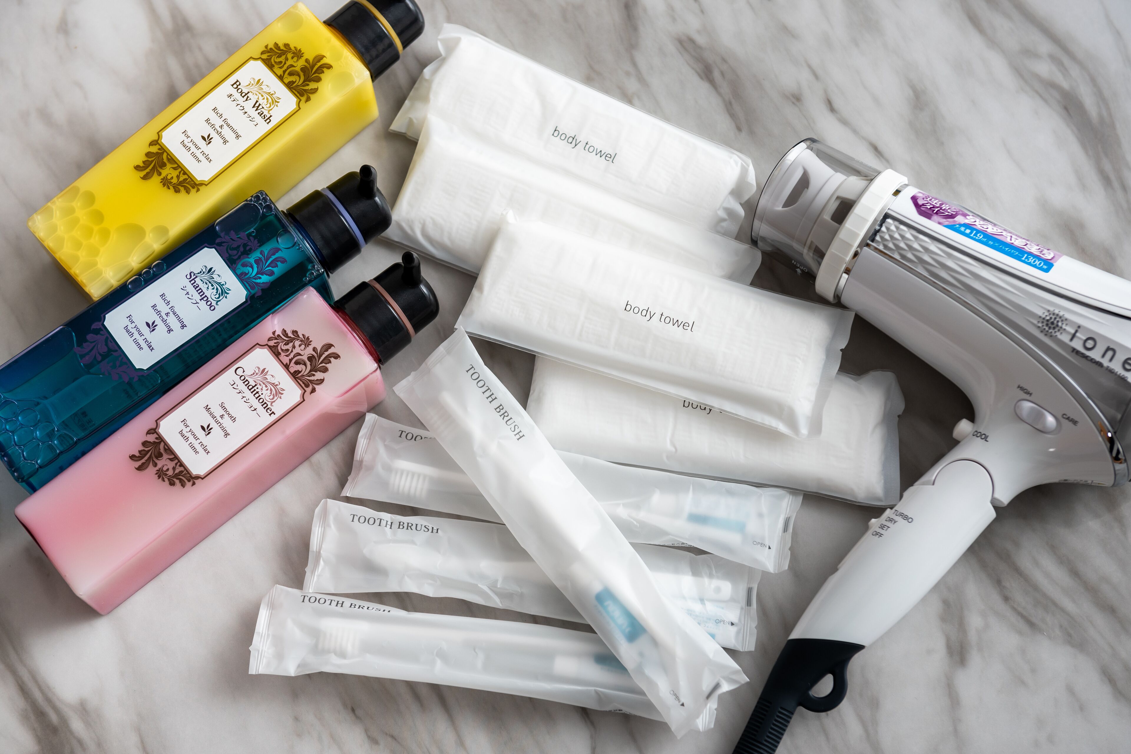 Variedad de artículos de aseo, incluyendo gel de baño, champú, acondicionador, toalla de baño y cepillos de dientes, sobre una superficie de mármol.