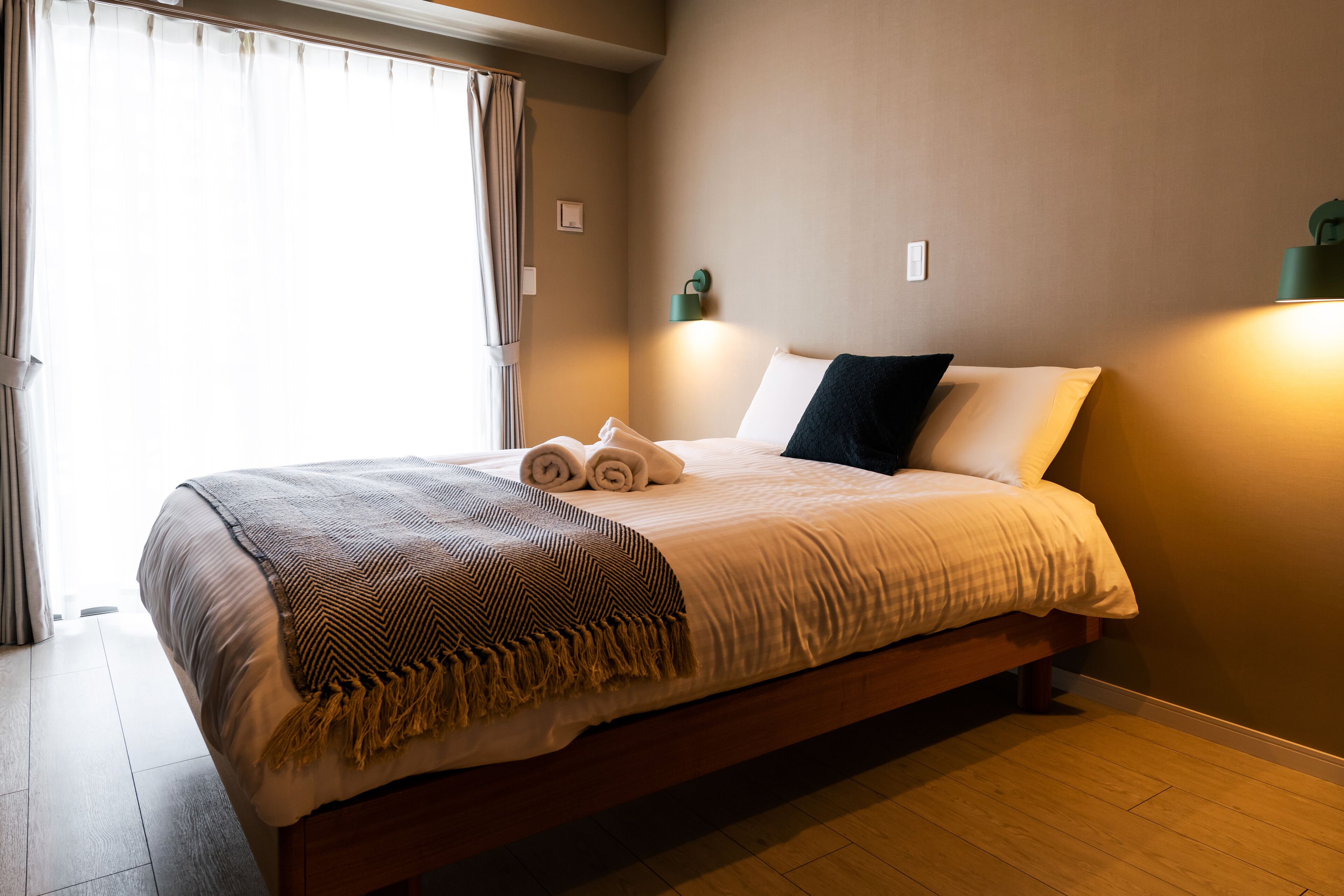 A neatly made bed with a blanket and rolled towels, a bedside lamp, and a window with curtains.