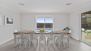 Een eetgedeelte met een houten tafel en witte stoelen, een groot raam met uitzicht op een zwembad en ingelijste foto's aan de muur.