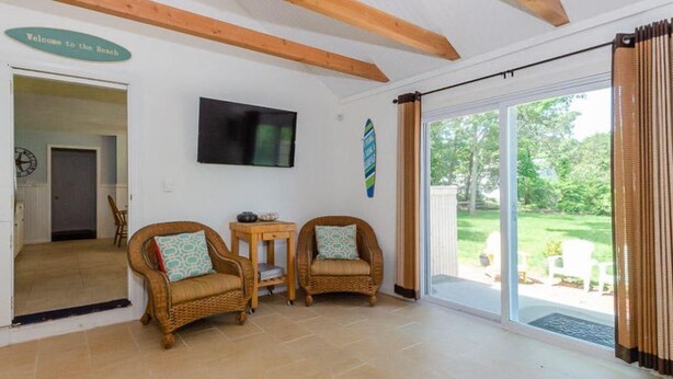 A room with wicker chairs, a small table, a flat-screen TV, and a sliding glass door leading to a garden.