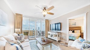 Uma sala de estar ampla com um sofá grande, uma mesa de centro e uma televisão. Há uma varanda com vista para a cidade.