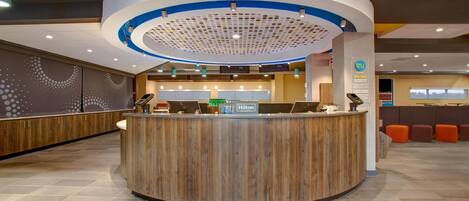 A modern hotel reception area with a curved wooden reception desk, multiple computer monitors, and a circular ceiling design.
