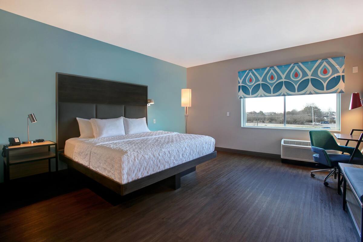 A hotel room with a large bed, a desk, a chair, a lamp, and a window with a view.