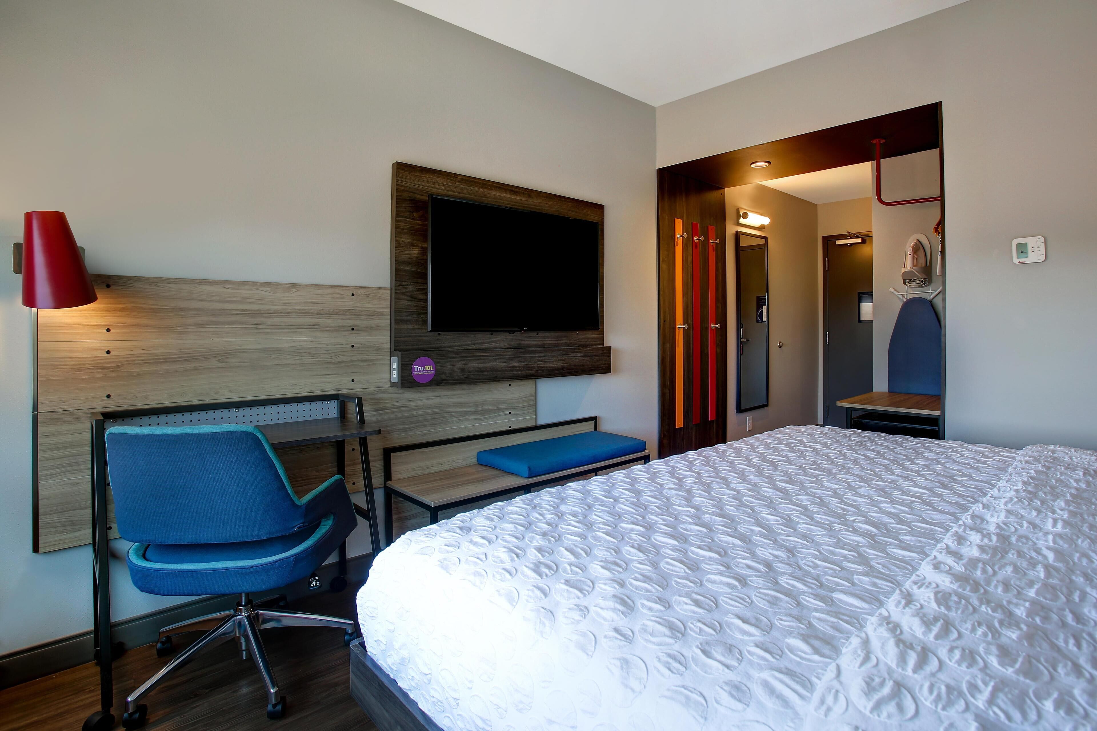 Habitación de hotel con cama, escritorio y silla, televisor montado en la pared y una lámpara roja.
