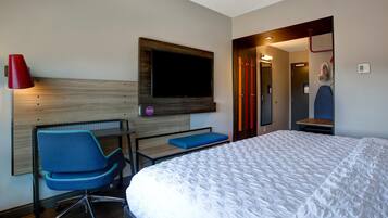 Habitación de hotel con cama, escritorio y silla, televisor montado en la pared y una lámpara roja.