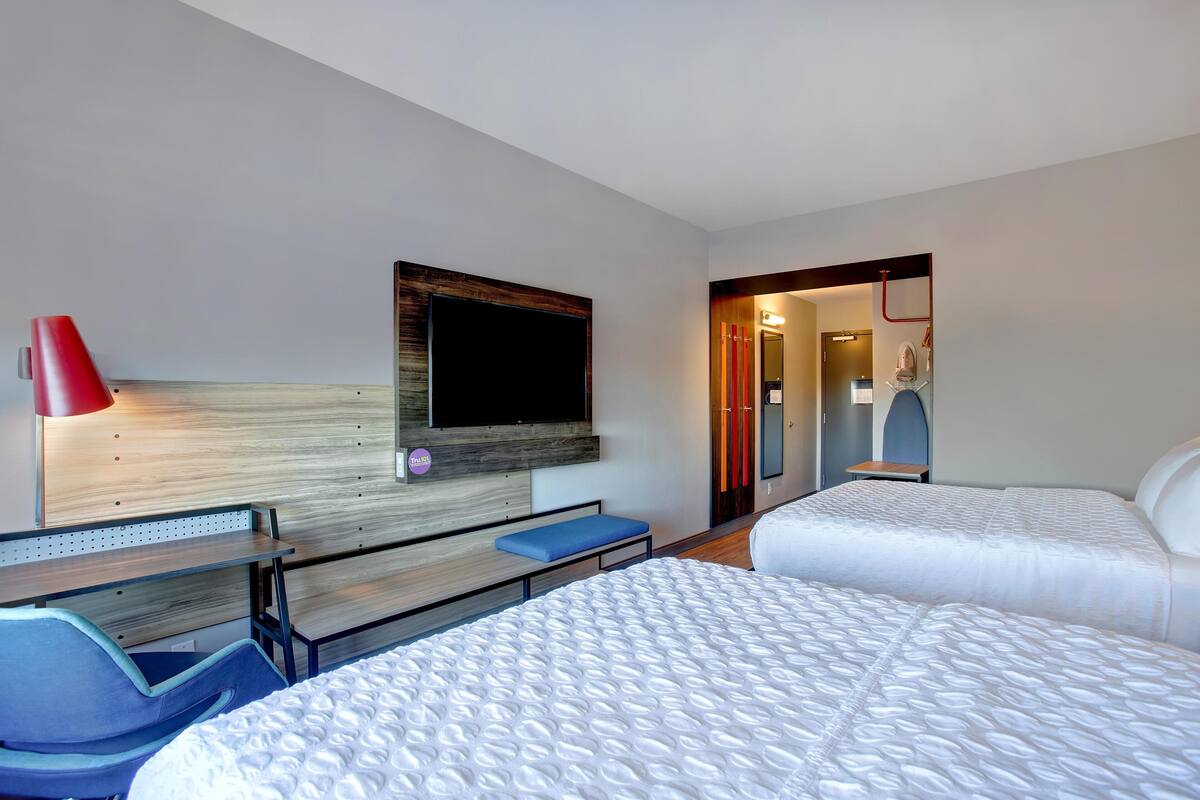 A hotel room with two beds, a desk, a chair, a TV mounted on the wall, and a red lamp.