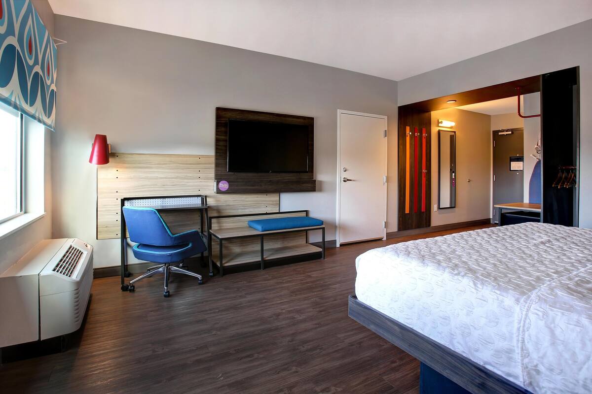 A hotel room with a bed, a desk with a chair, a TV mounted on the wall, a heater, and a door with a mirror.