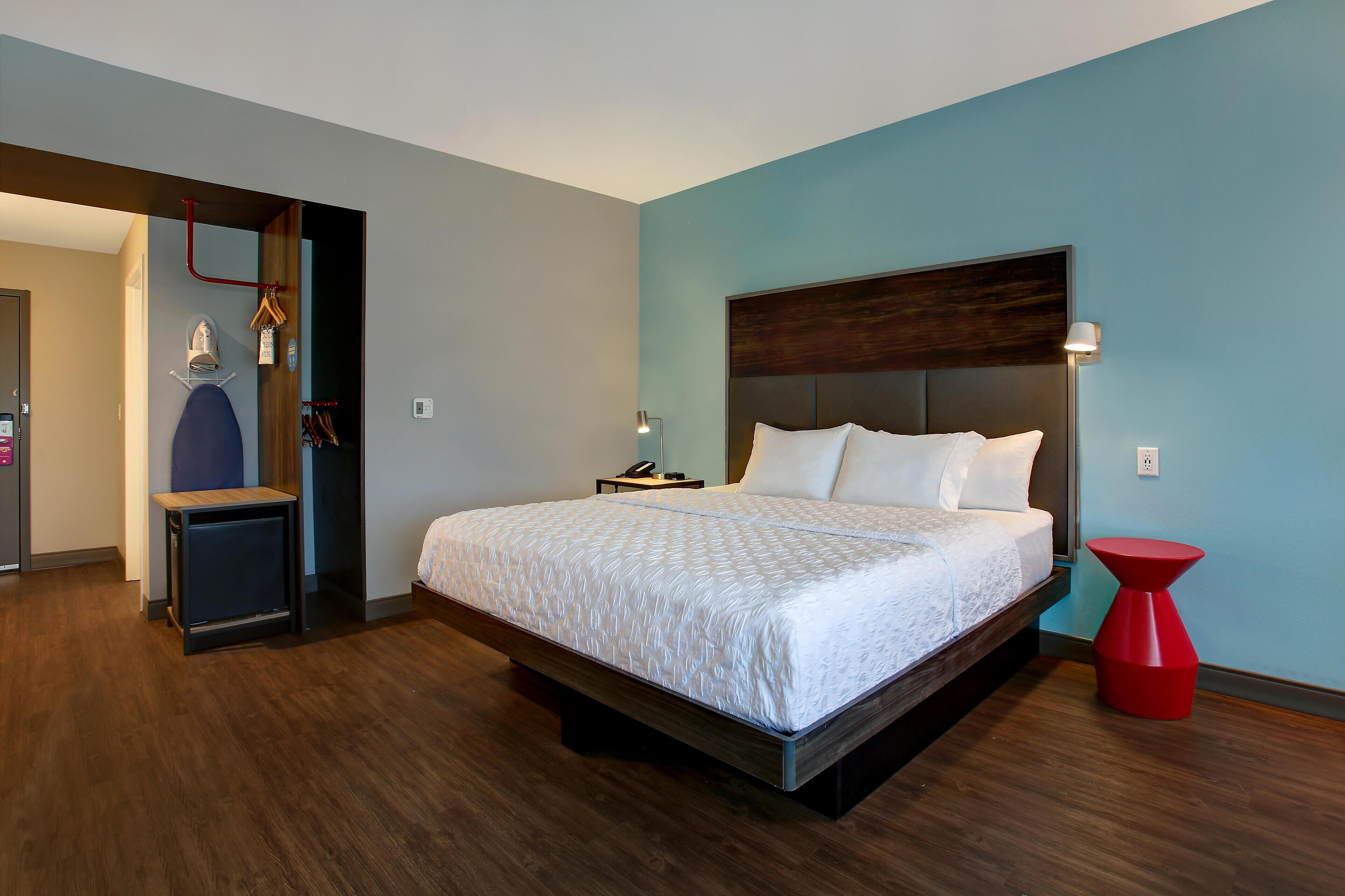 A modern hotel room with a large bed, a nightstand, a red side table, and a wooden floor.