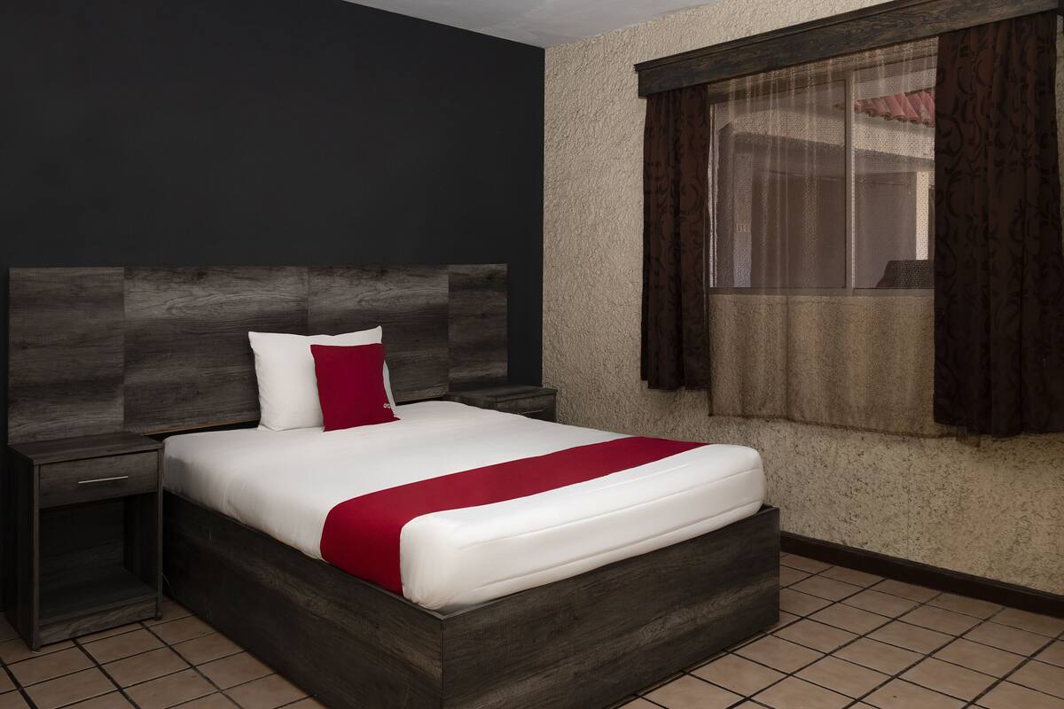 A neatly made bed with a white and red bedspread, a dark headboard, and a window with curtains.