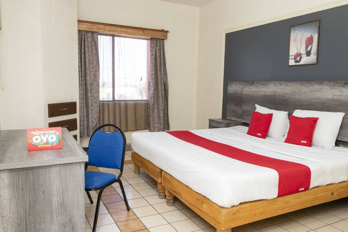 A hotel room with a wooden bed, a desk with a chair, a window with curtains, and a painting on the wall.