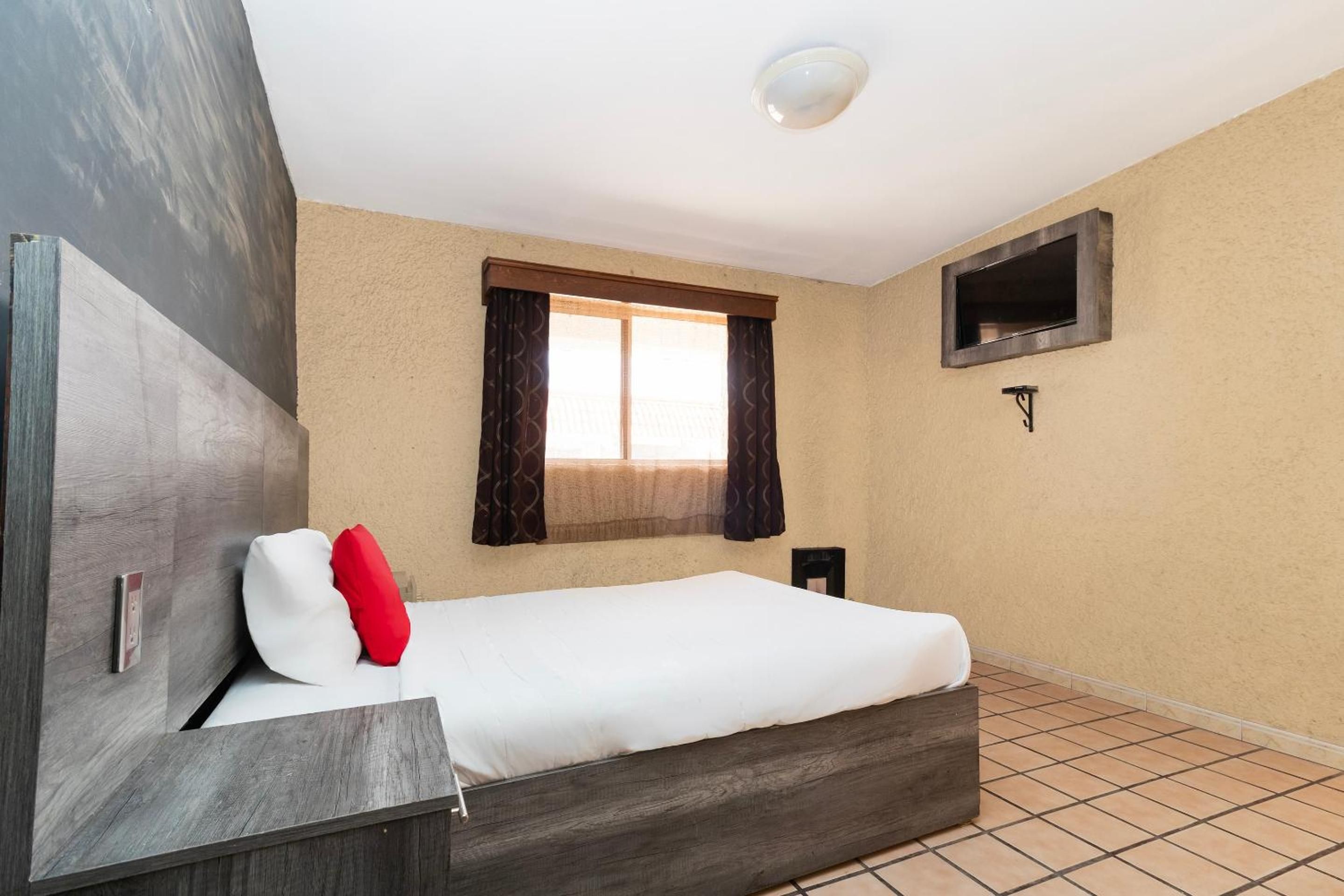 Un dormitorio con una cama, un televisor montado en la pared, una ventana con cortinas y piso de baldosas.