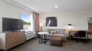 A hotel room with a flat-screen TV on a wooden dresser, a sofa with a side table, a desk with a chair, and a large window with curtains. - Staybridge Suites Atlanta Ne - Duluth by IHG (Duluth)