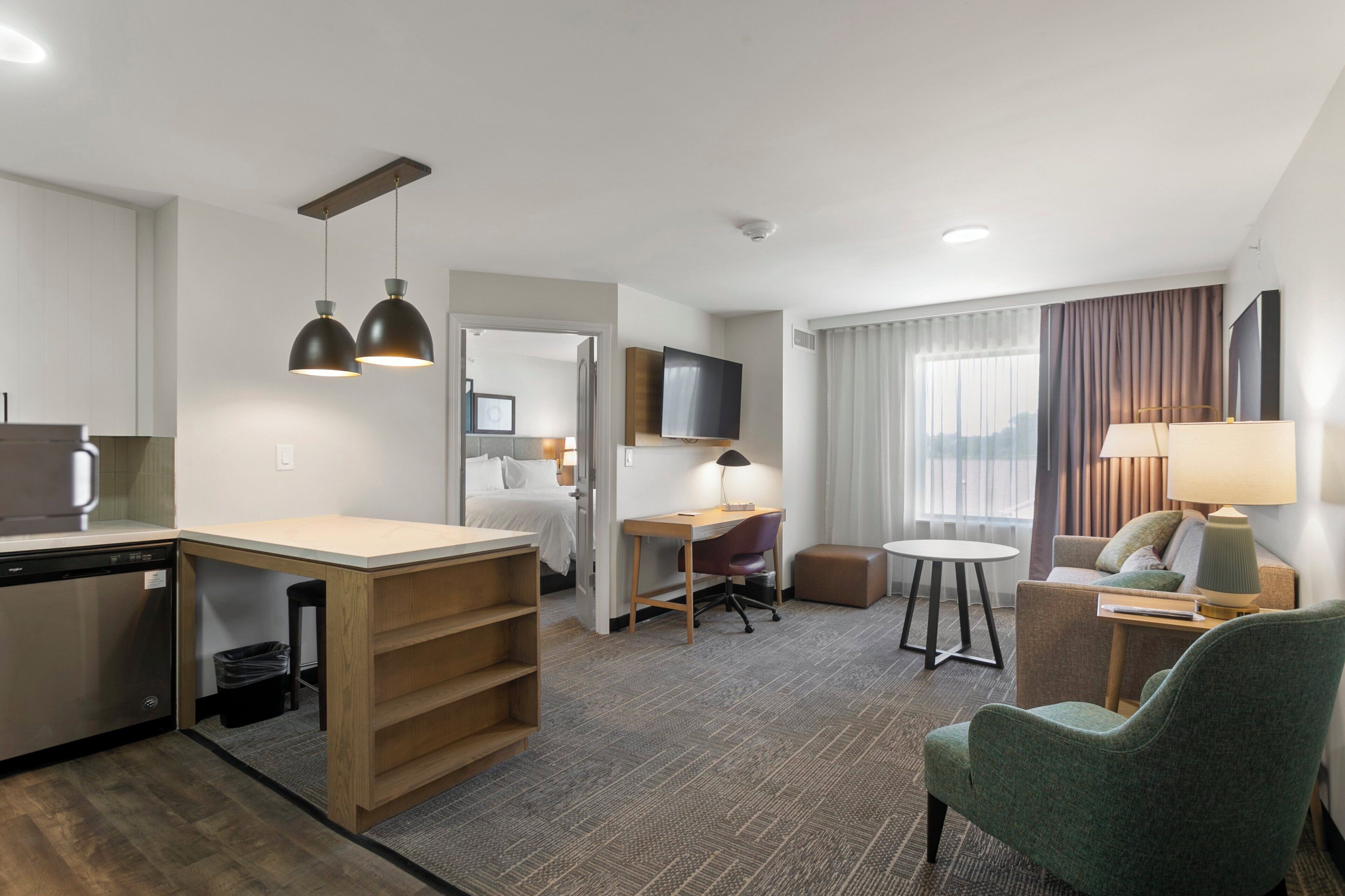 A hotel room with a kitchenette, a dining area, a seating area with a sofa and armchairs, a TV, and a window with curtains.