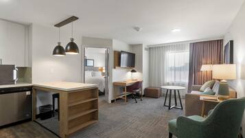 A hotel room with a kitchenette, a dining area, a seating area with a sofa and armchairs, a TV, and a window with curtains.