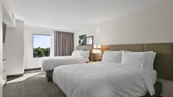 A hotel room with two beds, a large window with curtains, a bedside table with a lamp, and a view of greenery outside.