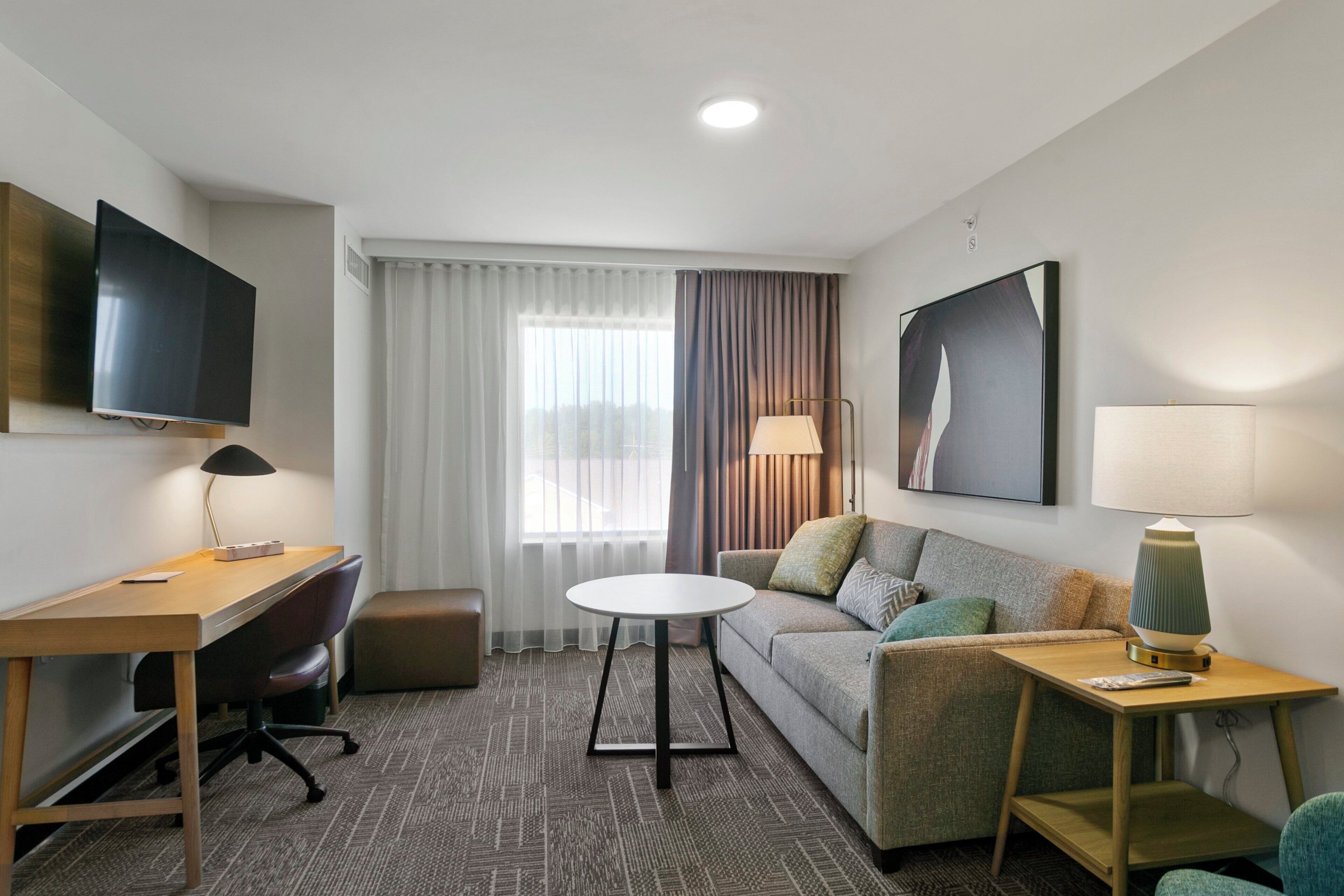 Habitación de hotel con escritorio, sofá, mesa redonda, televisor y ventana con cortinas.