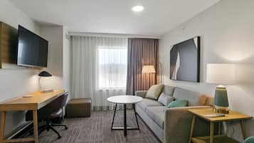 Habitación de hotel con escritorio, sofá, mesa redonda, televisor y ventana con cortinas.