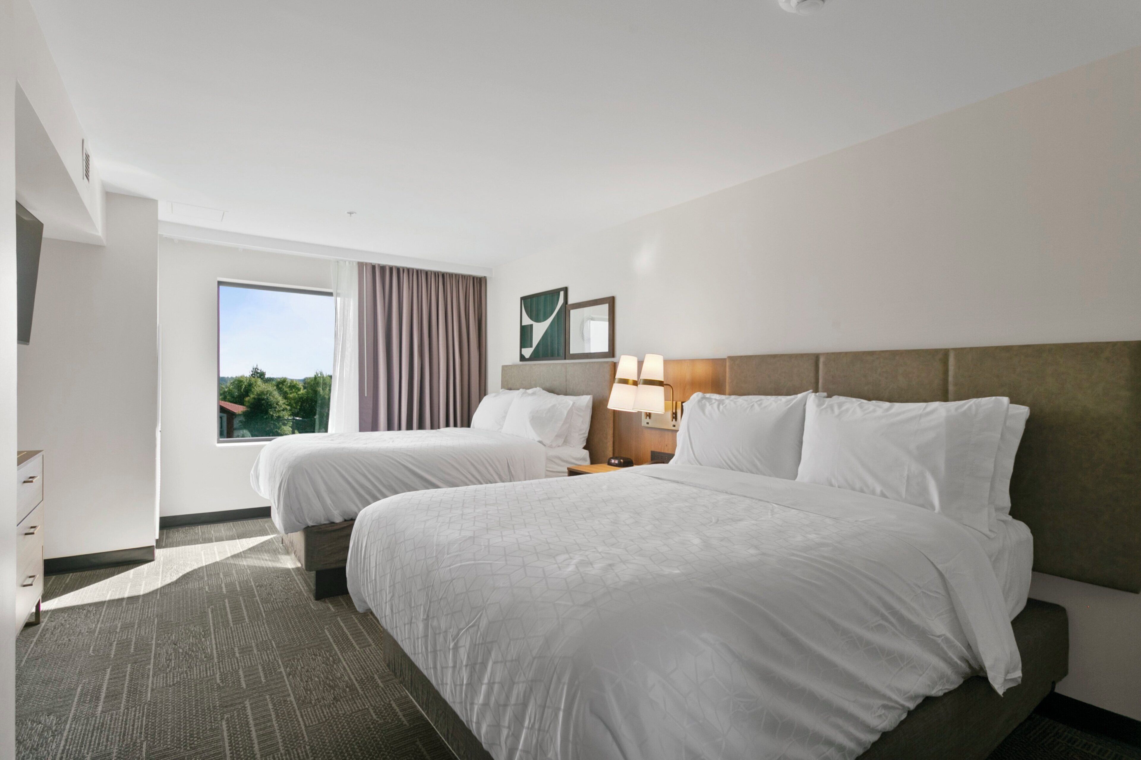 Habitación de hotel con dos camas, una ventana con cortinas, una mesita de noche con lámpara y un espejo montado en la pared.
