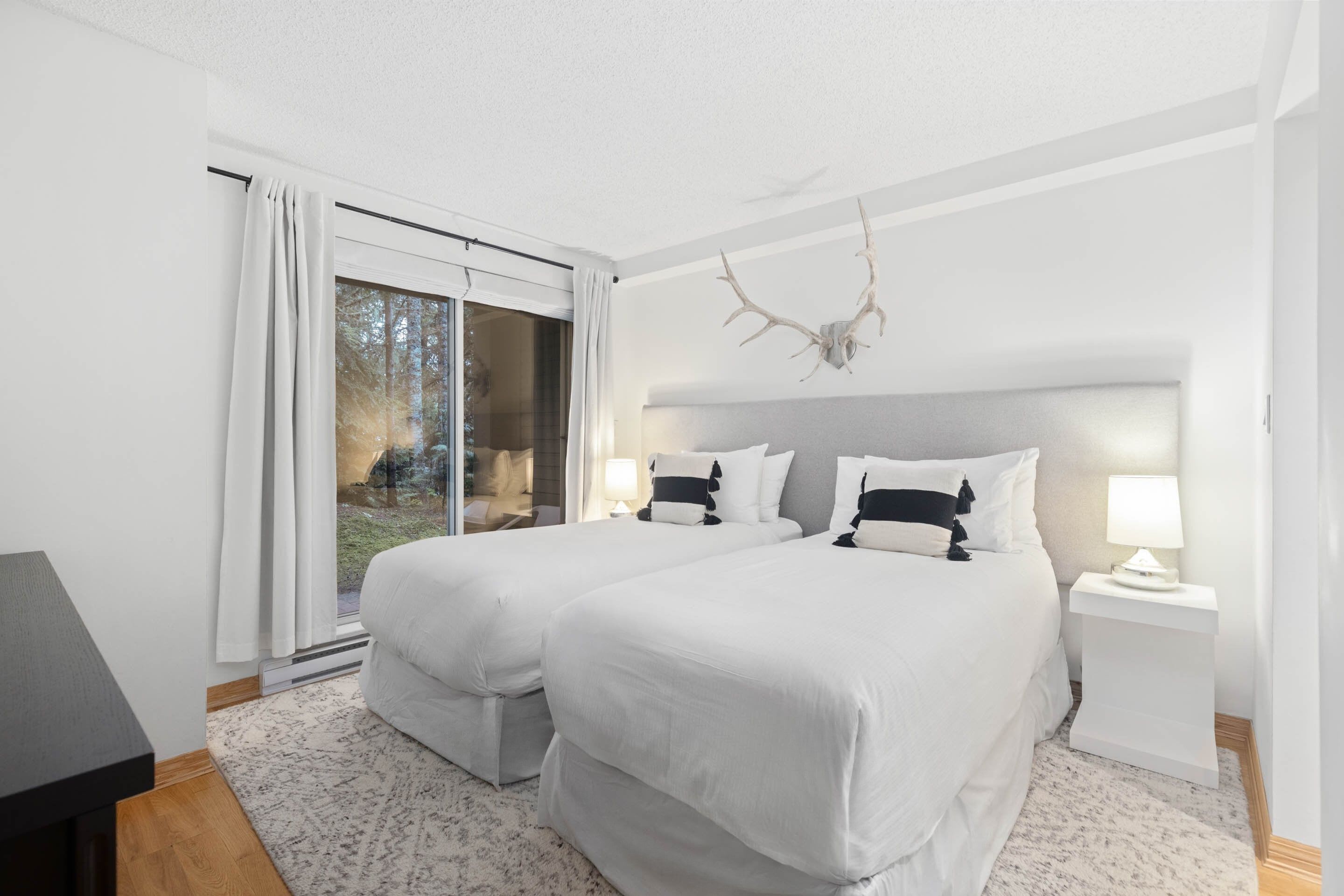 A modern hotel room with two beds, a large window with curtains, a deer antler decoration, and a small bedside table with a lamp.