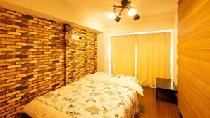 A bedroom with a brick wall, a wooden floor, a bed with a floral bedspread, a ceiling fan, and a window with curtains. - GrandTree Yokohama Station (Yokohama)