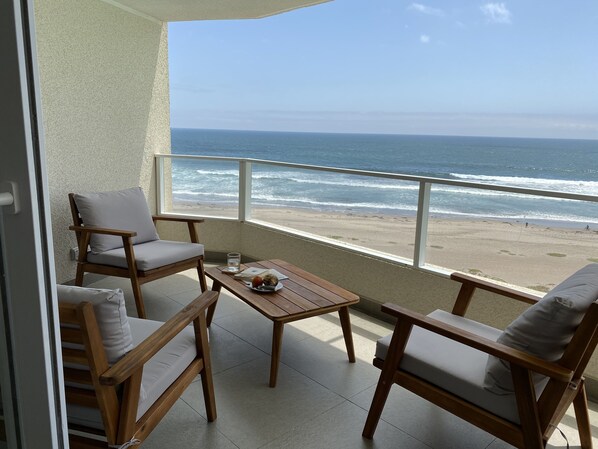 A balcony with a wooden table and two chairs, overlooking a beach with waves.