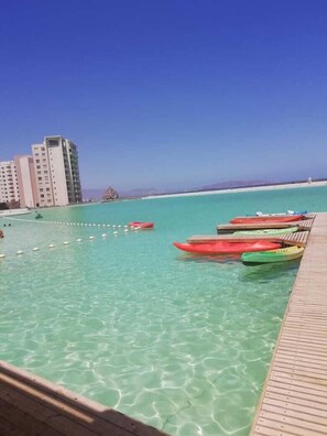A clear turquoise pool with kayaks lined up on a wooden deck, overlooking a beach and buildings.