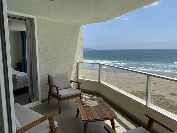 Un balcon avec une table et des chaises en bois, une vue sur la plage et un aperçu d’une pièce avec un lit et un rideau bleu.
