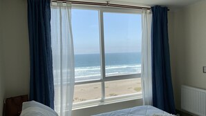 A bedroom with a large window overlooking a beach, a bed with white bedding, and a brown handbag on the floor. - Beachfront Spectacular New Apartment sleeps up to 8 - Laguna del Mar (La Serena)