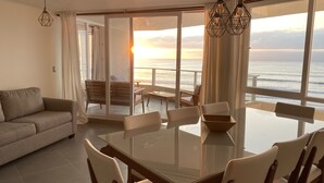 A modern living room with a glass dining table, comfortable seating, and a view of the ocean at sunset. - Beachfront Spectacular New Apartment sleeps up to 8 - Laguna del Mar (La Serena)