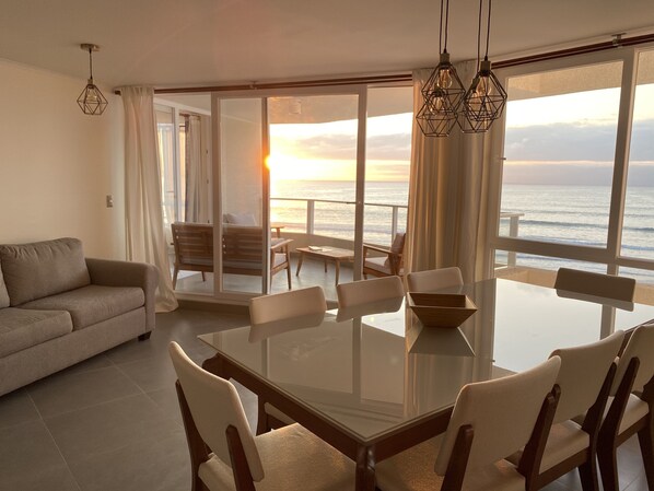 A modern living room with a glass dining table, comfortable seating, and a view of the ocean at sunset.