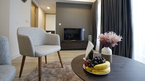 A modern living room with a grey armchair, a round coffee table with fruits and a bottle, a flat-screen TV, and a vase with flowers. - The First Stay Hotel (Phung Cong)