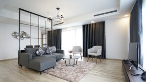 A modern living room with a grey sofa, a small round coffee table, a TV on a wooden stand, and a large window with curtains. - The First Stay Hotel (Phung Cong)