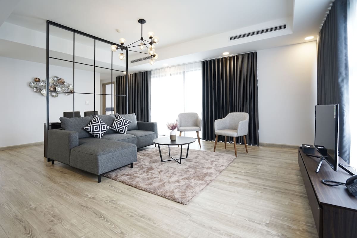 Uma sala de estar moderna com um sofá cinzento, uma mesa de centro redonda pequena, uma televisão sobre um suporte de madeira e uma janela grande com cortinas.