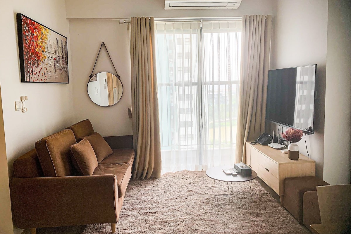 Uma sala de estar com sofá, televisão, mesa pequena, espelho e janela com cortinas.
