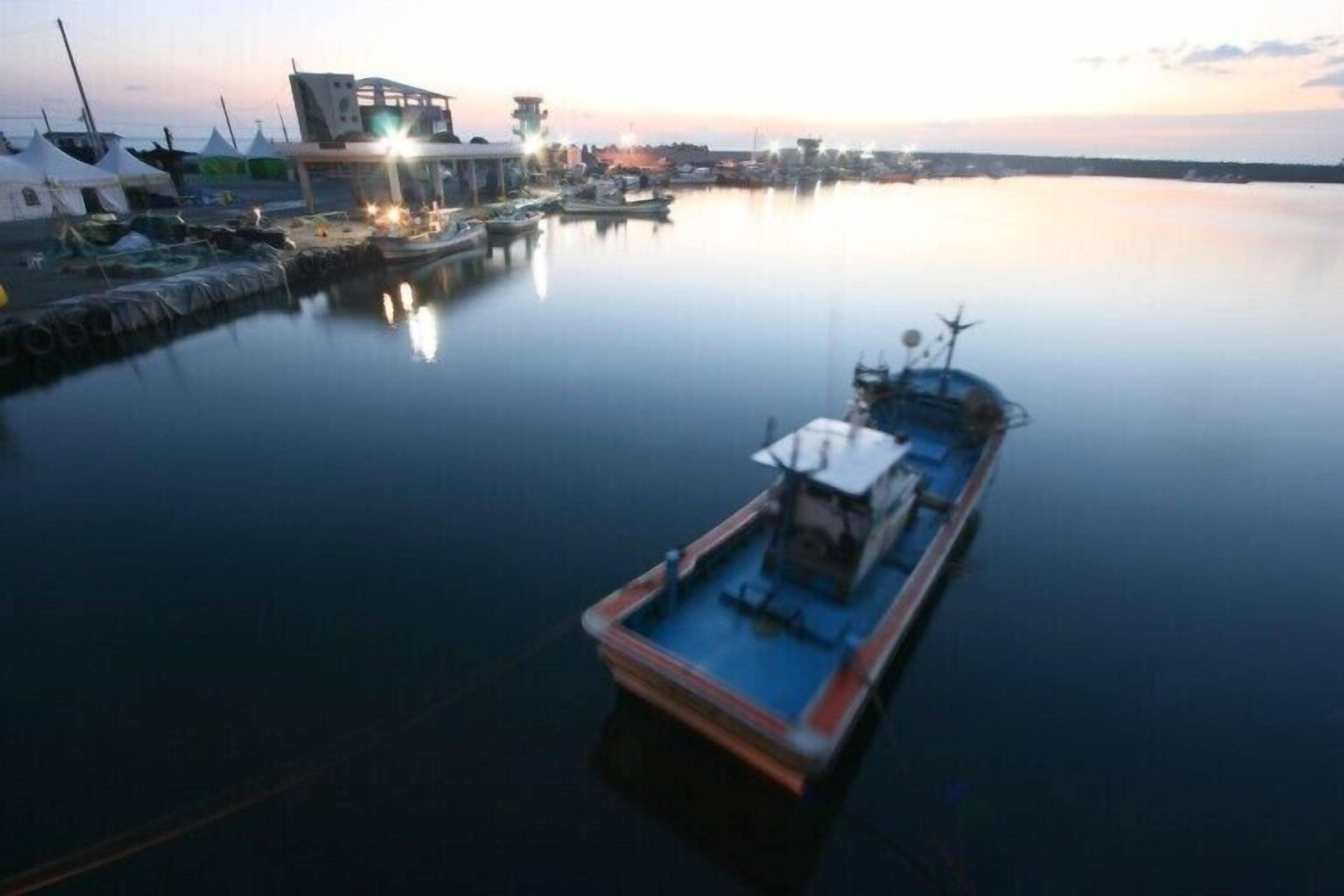A calm harbor with a docked boat, a building with lights, and tents on the shore.