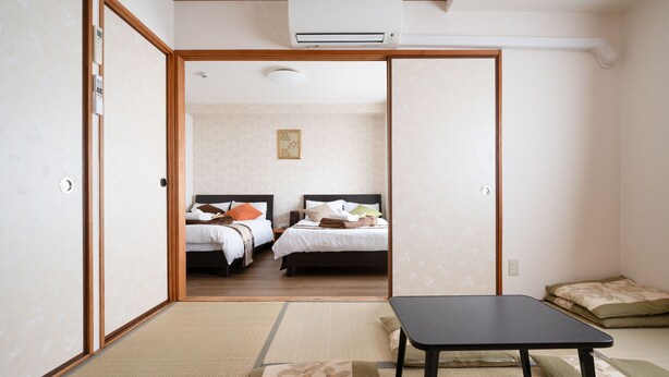 A room with two beds, a small table, and a futon. There is an air conditioning unit on the ceiling.