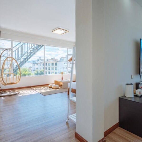 A spacious living room with a large window, a hanging chair, a sofa, and a flat-screen TV mounted on the wall.