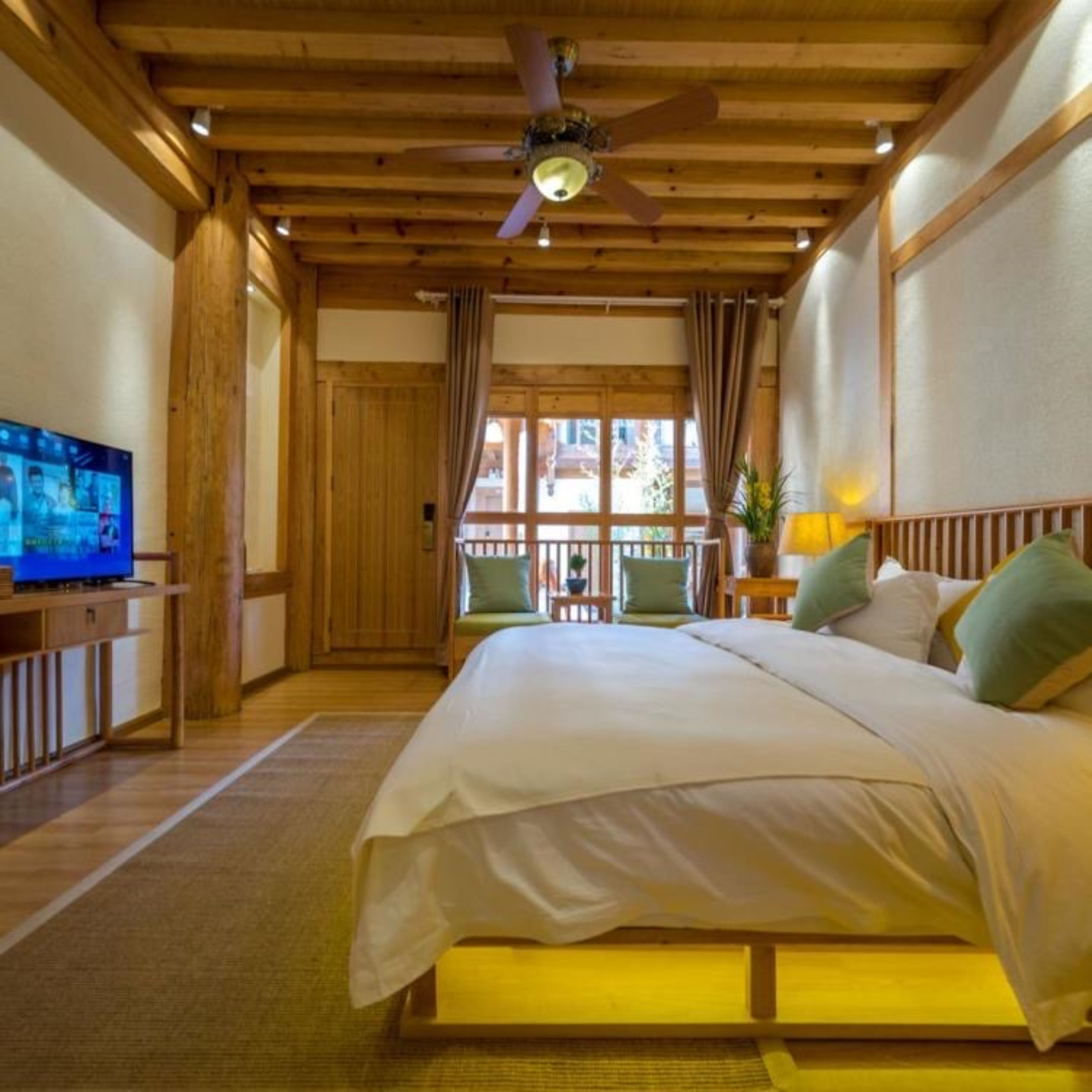 A bedroom with a large bed, wooden ceiling beams, a ceiling fan, a television on a wooden stand, and a window with curtains.