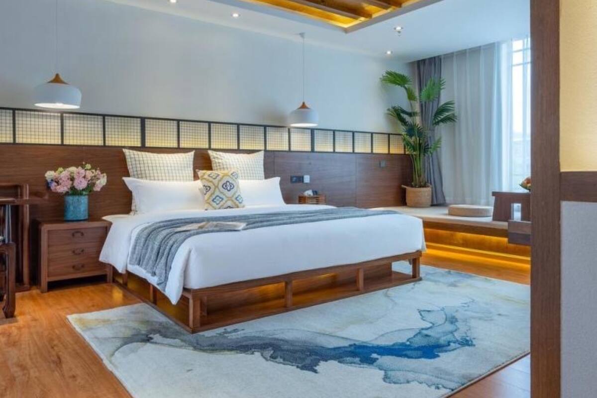 A modern bedroom with a wooden bed frame, a large window with curtains, a potted plant, and a nightstand with a lamp.