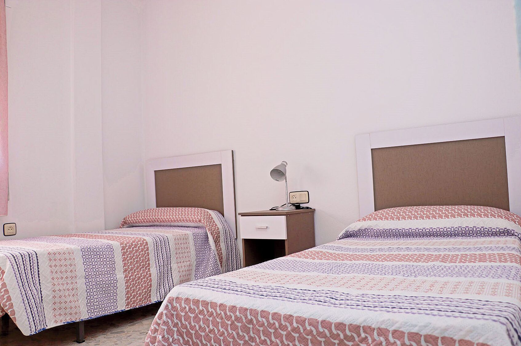A hotel room with two single beds, each with patterned bedding, a bedside table with a lamp, and a power outlet.