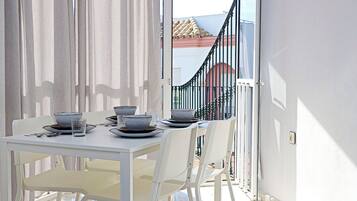 Un comedor con una mesa preparada para dos, sillas blancas y vista a un edificio de techos rojos a través de una puerta de balcón.