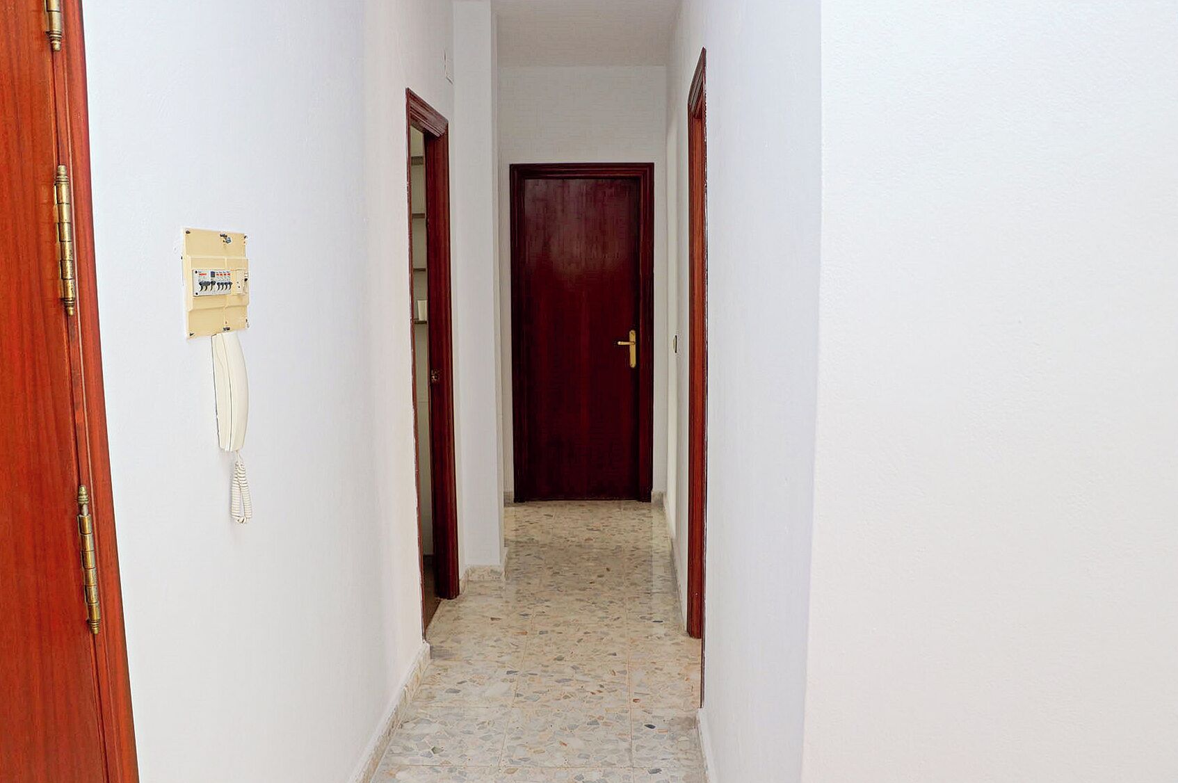A narrow hallway with a tiled floor, a wooden door at the end, and a wall-mounted telephone.