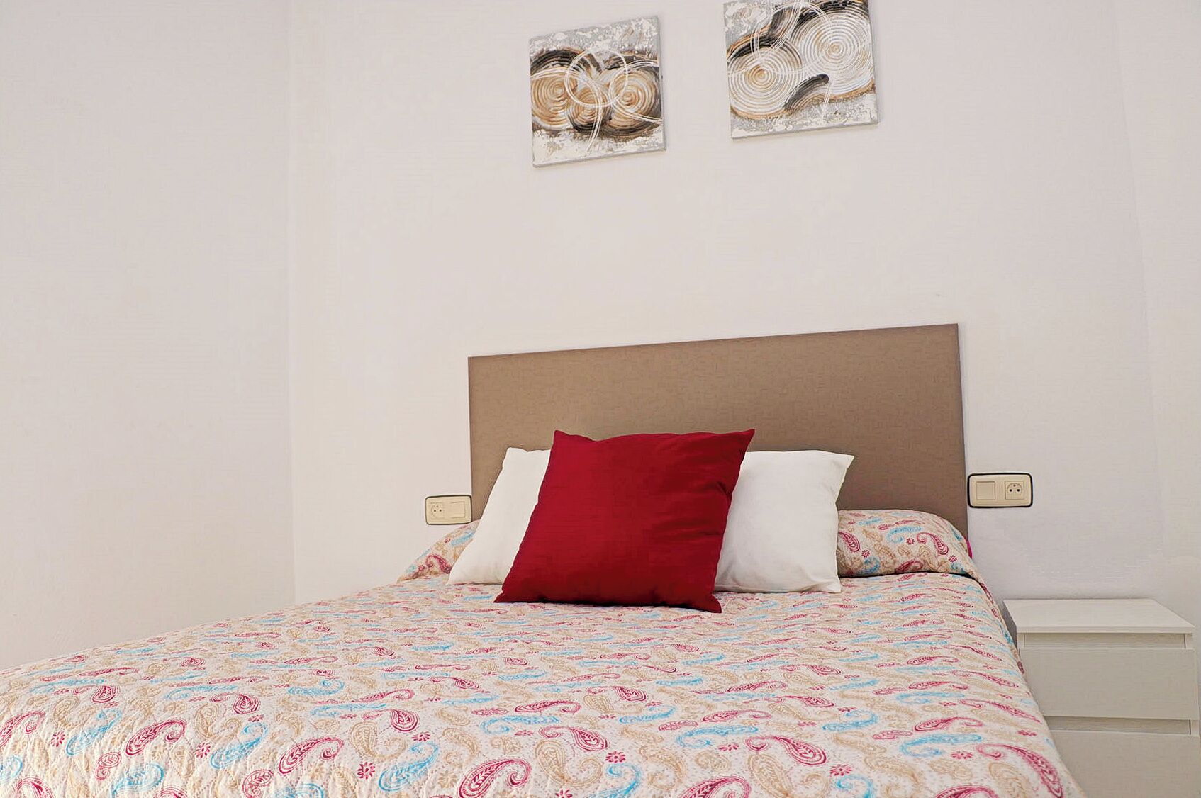 Een bed met een bedsprei met dessin, twee kussens, een houten hoofdbord, nachtkastjes en twee aan de muur bevestigde kunstwerken.